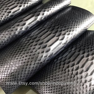 May include: Close-up of black and silver python leather. The leather has a textured, scaly pattern. The leather is folded in a stack, showing the different shades of black and silver.