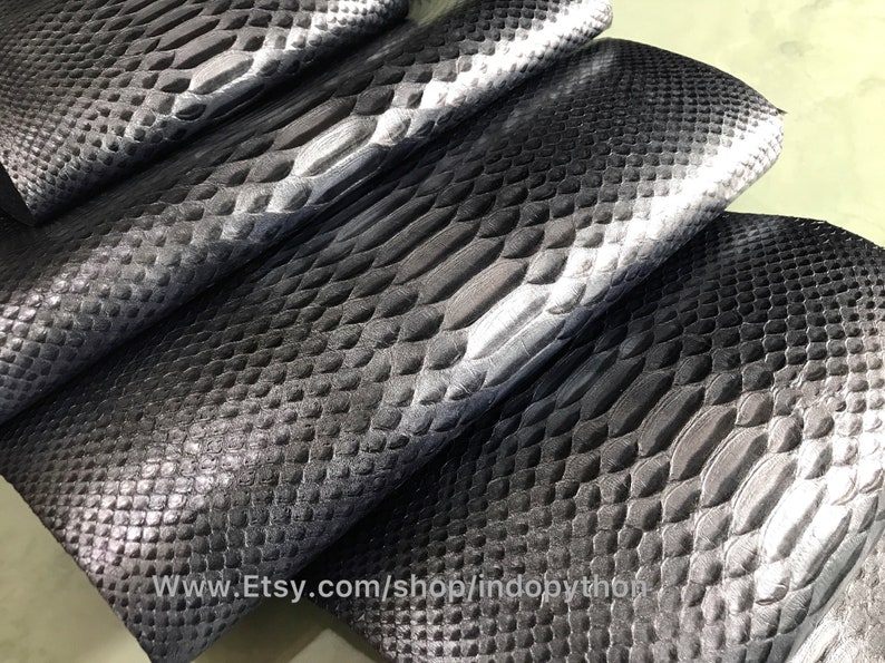 May include: Close-up of black and silver python leather. The leather has a textured, scaly pattern. The leather is folded in a stack, showing the different shades of black and silver.