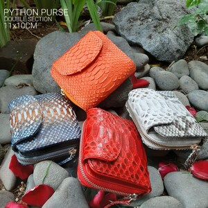 K&ouml;nnte beinhalten: Vier verschiedene farbige Python-Leder-Geldb&ouml;rsen mit Rei&szlig;verschl&uuml;ssen. Die Geldb&ouml;rsen liegen auf einem Bett aus kleinen grauen Steinen und Rosenbl&uuml;ten. Die Geldb&ouml;rsen sind 11x10 cm gro&szlig;.