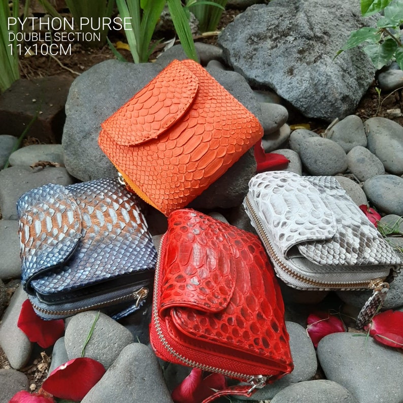 K&ouml;nnte beinhalten: Vier verschiedene farbige Python-Leder-Geldb&ouml;rsen mit Rei&szlig;verschl&uuml;ssen. Die Geldb&ouml;rsen liegen auf einem Bett aus kleinen grauen Steinen und Rosenbl&uuml;ten. Die Geldb&ouml;rsen sind 11x10 cm gro&szlig;.