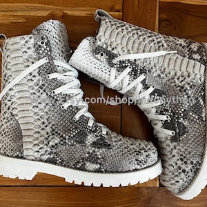 May include: A pair of white and gray snake skin boots with white laces. The boots have a white sole and a round toe.