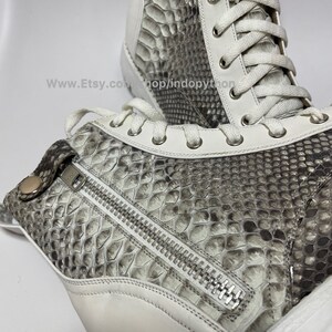 May include: White leather high-top sneakers with a snakeskin pattern and a zipper detail on the side. The laces are white and the sole is white.