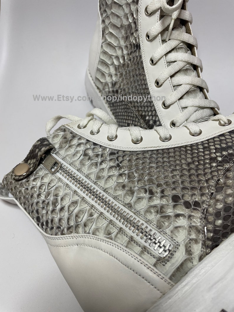 May include: White leather high-top sneakers with a snakeskin pattern and a zipper detail on the side. The laces are white and the sole is white.