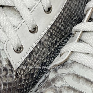 May include: Close-up of a pair of white leather sneakers with grey python skin detailing. The laces are white and the eyelets are silver. The shoes have a textured surface.
