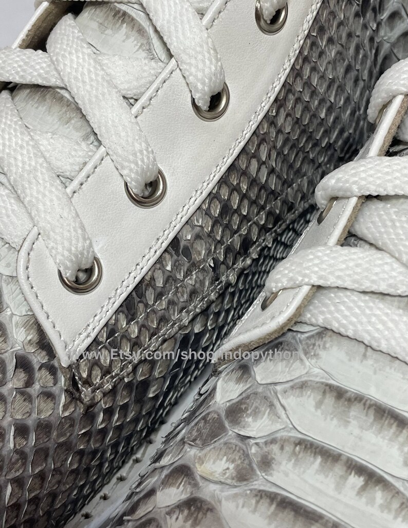 May include: Close-up of a pair of white leather sneakers with grey python skin detailing. The laces are white and the eyelets are silver. The shoes have a textured surface.
