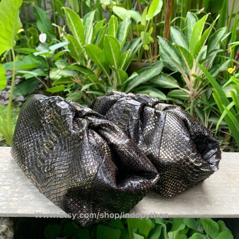K&ouml;nnte beinhalten: Zwei schwarze und goldene Clutch-Taschen aus Python-Leder mit gerafftem, ger&uuml;schtem Design. Die Taschen liegen auf einer grauen Oberfl&auml;che mit gr&uuml;nem Laub im Hintergrund.