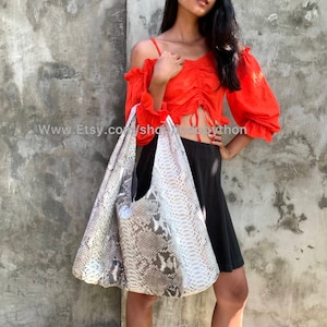 May include: A white and gray snakeskin tote bag with a long strap. The bag is being held by a person wearing a red off-the-shoulder top and a black skirt.