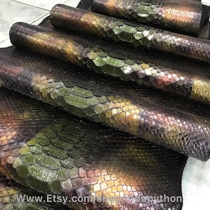 Puede incluir: Varias piezas de cuero de pitón iridiscente verde, marrón y dorado. El cuero tiene una textura escamosa y está dispuesto en una pila.