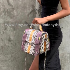 May include: A purple, white, and brown snakeskin handbag with a gold chain strap and a gold clasp. The bag has a top handle and a flap closure.