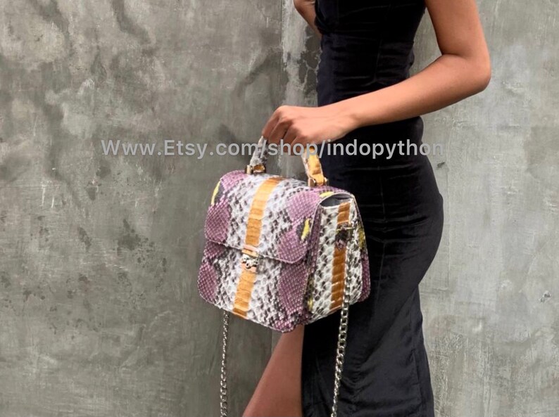 May include: A purple, white, and brown snakeskin handbag with a gold chain strap and a gold clasp. The bag has a top handle and a flap closure.