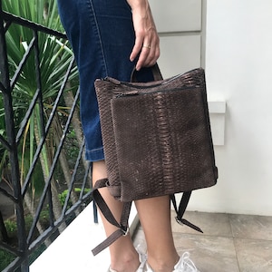 K&ouml;nnte beinhalten: Ein brauner Schlangenleder-Rucksack mit Rei&szlig;verschluss und verstellbaren Tr&auml;gern. Der Rucksack wird von einer Person getragen, die einen Jeansrock und wei&szlig;e Turnschuhe tr&auml;gt.