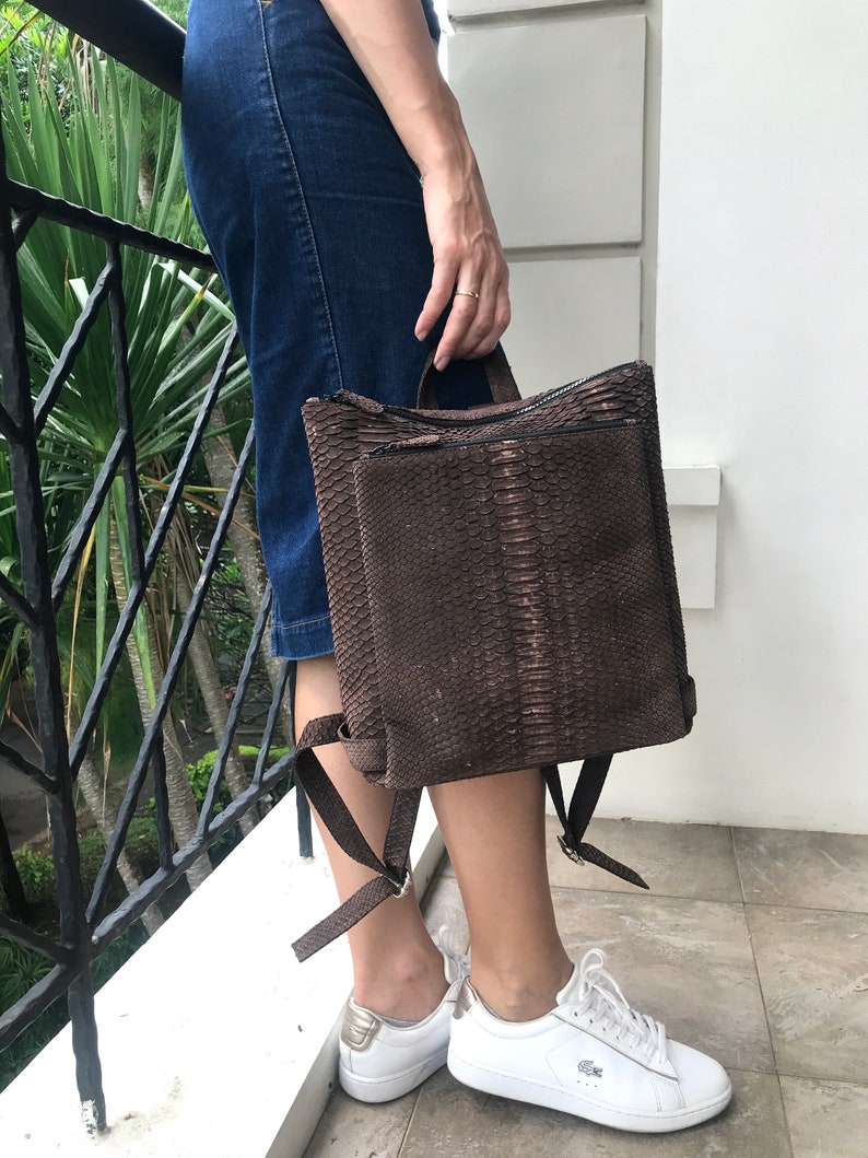 K&ouml;nnte beinhalten: Ein brauner Schlangenleder-Rucksack mit Rei&szlig;verschluss und verstellbaren Tr&auml;gern. Der Rucksack wird von einer Person getragen, die einen Jeansrock und wei&szlig;e Turnschuhe tr&auml;gt.