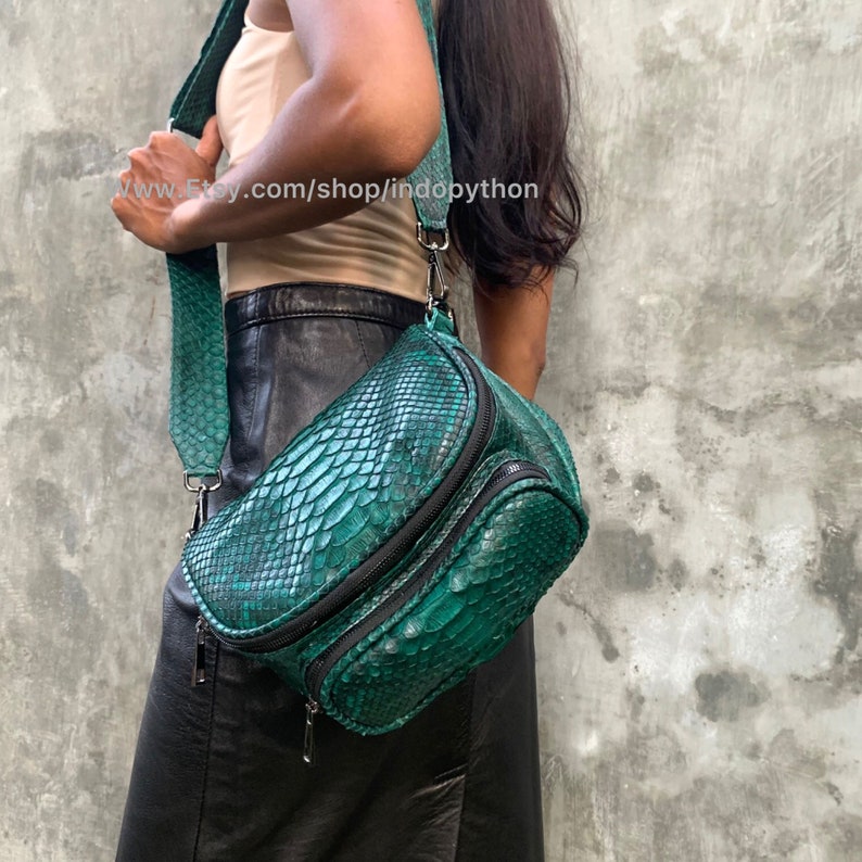 May include: A green snakeskin crossbody bag with a black zipper and strap. The bag has a unique curved shape and is made of genuine python leather.