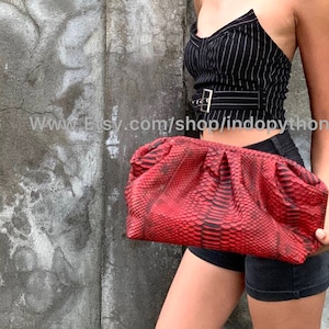 May include: A red python skin clutch bag with a gathered design. The bag is being held by a person wearing a black and white striped top and black shorts.