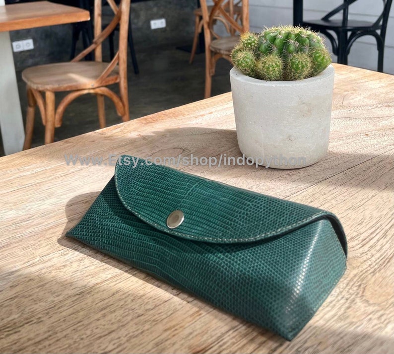 Op de afbeelding: Een donkergroene, getextureerde brillenkoker met een drukknoopsluiting. De koker is gemaakt van een materiaal dat op reptielenhuid lijkt. De koker is gesloten en ligt op een houten oppervlak. Een kleine cactus in een betonnen pot staat op de achtergrond.
