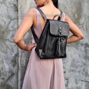 May include: Black leather backpack with a snakeskin pattern and a tassel. The backpack has a flap closure with a silver buckle and a zipped pocket on the front.