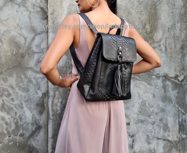 May include: Black leather backpack with a snakeskin pattern and a tassel. The backpack has a flap closure with a silver buckle and a zipped pocket on the front.