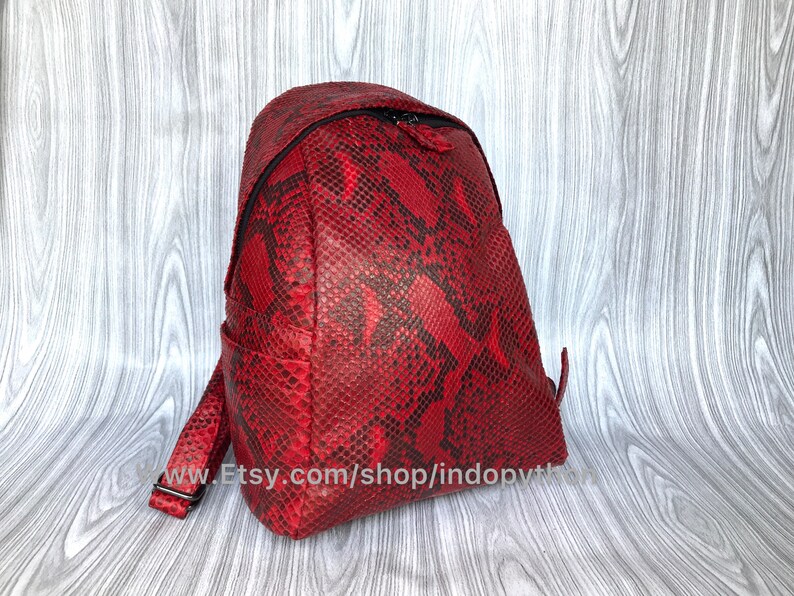 May include: A red snakeskin backpack with a black zipper and adjustable straps. The backpack is made of genuine leather and has a unique, textured design.