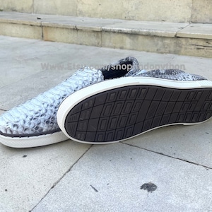 K&ouml;nnte beinhalten: Ein Paar graue Slipper aus Schlangenleder mit wei&szlig;en Sohlen. Die Schuhe haben ein strukturiertes Muster und sind von der Seite zu sehen.