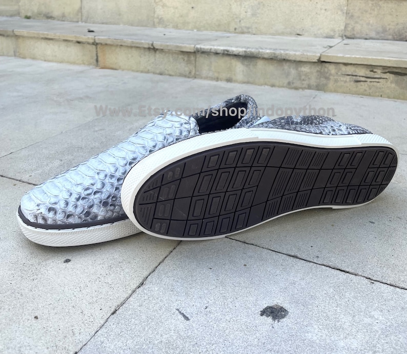 K&ouml;nnte beinhalten: Ein Paar graue Slipper aus Schlangenleder mit wei&szlig;en Sohlen. Die Schuhe haben ein strukturiertes Muster und sind von der Seite zu sehen.