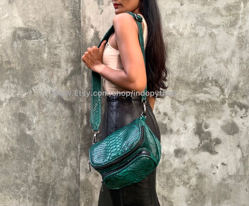 May include: A green snakeskin crossbody bag with a black zipper and strap. The bag has a unique, curved shape and is made of genuine leather.