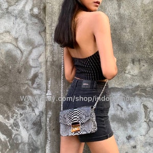 May include: A black and white snakeskin crossbody bag with a silver chain strap. The bag has a flap closure with a gold buckle. The bag is worn by a person wearing a black and white striped top and black denim shorts.