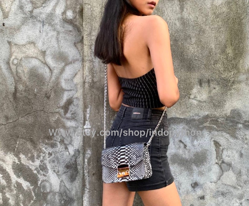 May include: A black and white snakeskin crossbody bag with a silver chain strap. The bag has a flap closure with a gold buckle. The bag is worn by a person wearing a black and white striped top and black denim shorts.