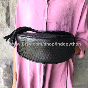 May include: A black round handbag made of python skin with a tassel. The bag has a long strap and is held by a person wearing a pink shirt.