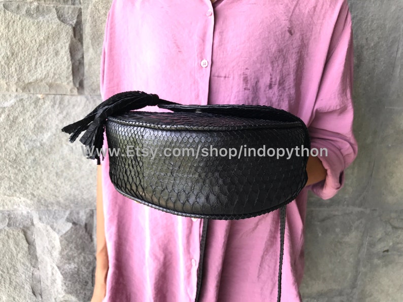 May include: A black round handbag made of python skin with a tassel. The bag has a long strap and is held by a person wearing a pink shirt.