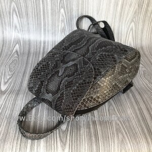 K&ouml;nnte beinhalten: Ein kleiner grauer und brauner Schlangenleder-Rucksack mit Rei&szlig;verschluss. Der Rucksack hat eine Klappe mit Schnallenverschluss und ein kleines Fach auf der Vorderseite.