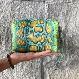 Peut inclure: Une pochette rectangulaire avec un motif peau de serpent jaune, bleu et vert. La pochette est dotée d'une dragonne et d'une fermeture éclair. Le nom de la boutique Etsy est visible.
