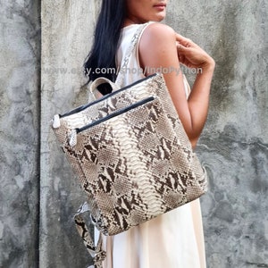 Könnte beinhalten: Ein weißer und brauner Schlangenleder-Rucksack mit schwarzen Reißverschlüssen. Der Rucksack hat einen oberen Griff und verstellbare Träger.