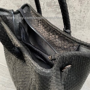 Pode incluir: Uma bolsa de m&atilde;o de couro preto com padr&atilde;o de pele de cobra e fecho de z&iacute;per. A bolsa tem duas al&ccedil;as superiores e um detalhe de metal prateado no lado.