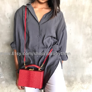 Puede incluir: Un bolso de mano rojo de piel de serpiente con cierre dorado y correa roja. El bolso lo lleva una persona que lleva una camisa gris y pantalones blancos.