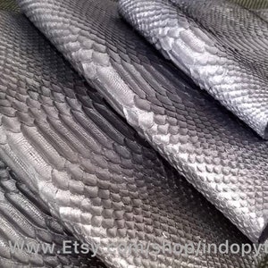 以下が含まれることがあります： 銀色のパイソン革が4枚、重ねられています。革は、質感のある鱗模様をしています。革は半分に折り畳まれており、端が確認できます。革は緑色の背景の上に置かれています。 www.Etsy.com/shop/indopython