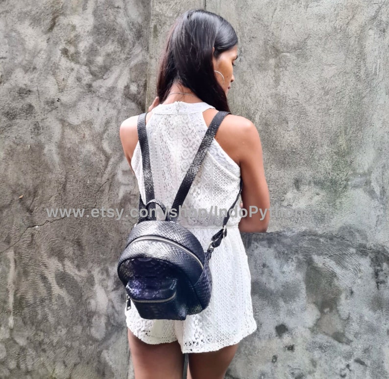 May include: A black leather backpack with a snakeskin pattern. The backpack has a zippered closure and adjustable straps. The backpack is being worn by a person in a white dress.