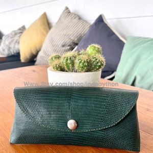 Op de afbeelding: Een donkergroene, getextureerde brillenkoker met een klepsluiting en een zilveren knoop. De rechthoekige koker, vermoedelijk van leer, ligt op een houten tafel. Achter de koker staat een kleine cactus in een witte pot.