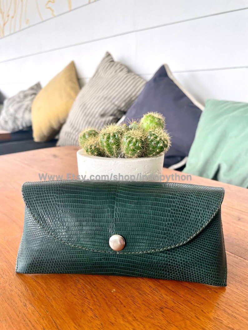 Op de afbeelding: Een donkergroene, getextureerde brillenkoker met een klepsluiting en een zilveren knoop. De rechthoekige koker, vermoedelijk van leer, ligt op een houten tafel. Achter de koker staat een kleine cactus in een witte pot.