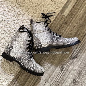 Puede incluir: Un par de botas de piel de serpiente negra y blanca con cordones. Las botas tienen una suela gruesa y están hechas de un material texturizado.