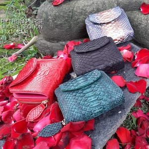 K&ouml;nnte beinhalten: Vier verschiedene farbige Python-Ledergeldb&ouml;rsen mit einem Doppelfach-Design. Die Geldb&ouml;rsen sind 11 x 10 cm gro&szlig; und liegen auf einem Bett aus roten Rosenbl&auml;ttern.