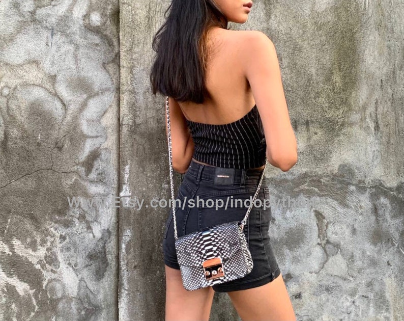 May include: A black and white snakeskin crossbody bag with a silver chain strap and a gold clasp. The bag is worn by a person wearing a black and white striped top and black denim shorts.