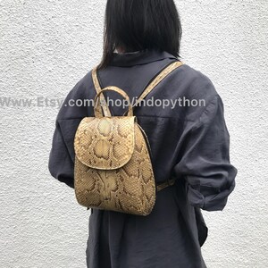 Puede incluir: Una peque&ntilde;a mochila de piel de serpiente marr&oacute;n con cierre de solapa. La mochila la lleva puesta una persona con el pelo negro y una camisa gris. El texto "www.Etsy.com/shop/lindopython" es visible en la imagen.