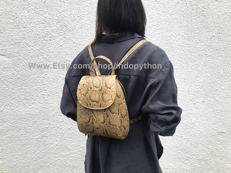 Puede incluir: Una peque&ntilde;a mochila de piel de serpiente marr&oacute;n con cierre de solapa. La mochila la lleva puesta una persona con el pelo negro y una camisa gris. El texto "www.Etsy.com/shop/lindopython" es visible en la imagen.