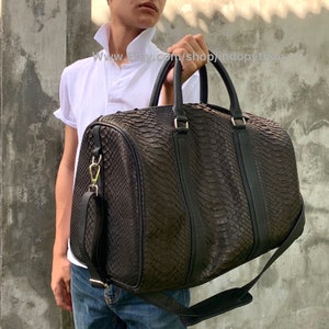 Puede incluir: Una bolsa de viaje de cuero negro con un patrón de piel de serpiente. La bolsa tiene un asa superior y una correa para el hombro.