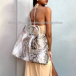 May include: A large, white and grey snakeskin tote bag with two top handles and a long shoulder strap. The bag is being worn by a person in a light beige dress.