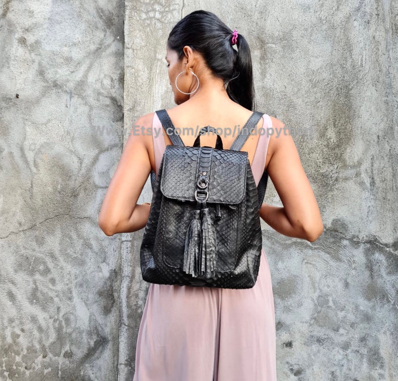 May include: Black leather backpack with a snakeskin pattern and a tassel. The backpack has a flap closure and a zipped pocket on the front.