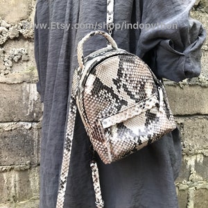 May include: A small, brown and white snakeskin backpack with a zip closure. The backpack has a long strap that can be adjusted to fit different heights.