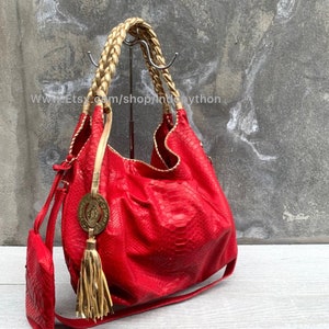 Può includere: Una borsa a mano in pelle di pitone rossa con manici intrecciati dorati e un pendente a nappa dorato. La borsa ha un emblema circolare dorato con un disegno.