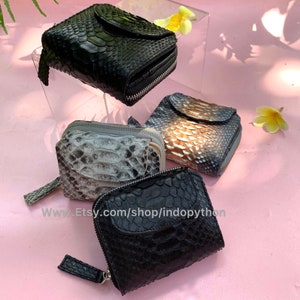 K&ouml;nnte beinhalten: Vier kleine Rei&szlig;verschlussportemonnaies in verschiedenen Farben aus Schlangenleder. Die Portemonnaies sind auf einer rosa Oberfl&auml;che angeordnet. Die Portemonnaies sind schwarz, grau, braun und blau.