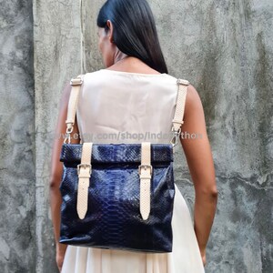 May include: A dark blue python leather backpack with cream-colored straps and buckles. The bag has a fold-over top and is worn by a person.